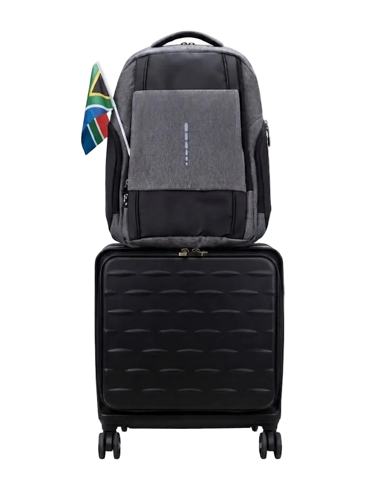 A gray and black backpack stacked on top of a black rolling suitcase against a plain white background, with a small South African flag hanging from the side of the backpack.
