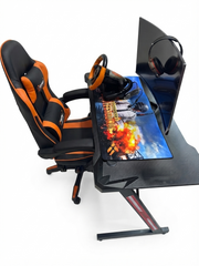 A gaming setup featuring a black and orange racing-style chair, a black desk with a large printed mouse pad, a steering wheel, and a monitor on a white background.